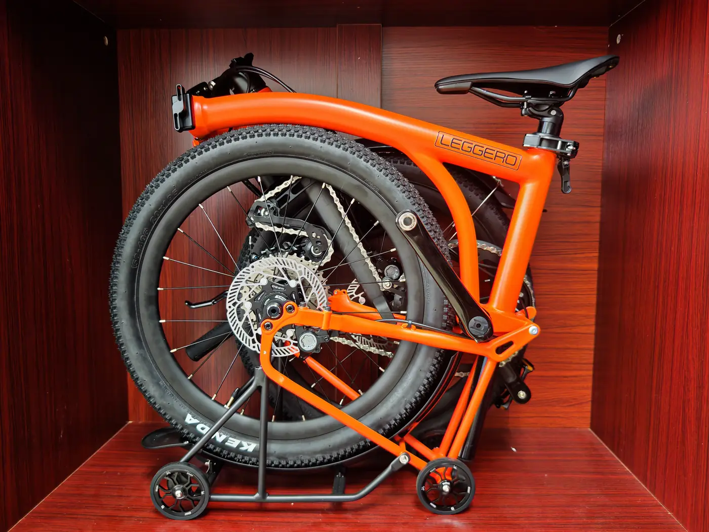 An orange Leggero Explorer GS with knobbly Kenda tyres, folded in a red wooden display cubby