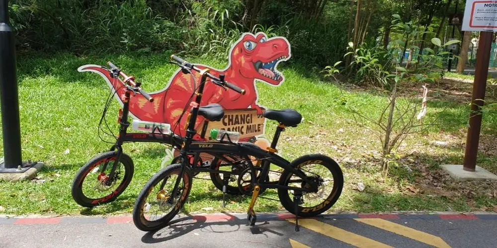 Two black folding bikes parked in front of a red T-rex sign on the Changi cycling path